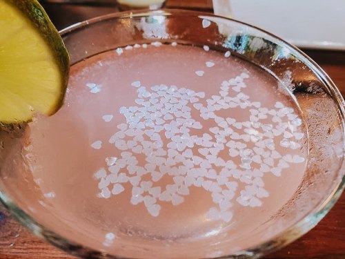 A close-up of a cocktail in a glass, garnished with a lime slice, featuring heart-shaped salt on the rim.