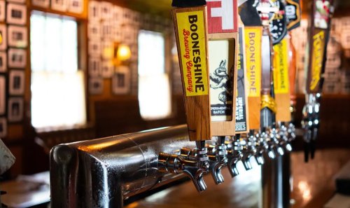Close-up of beer taps with various labels in a cozy bar setting, featuring Boone Shine prominently.