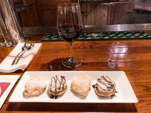 A plate with four desserts drizzled with chocolate, alongside a glass of red wine on a wooden bar.