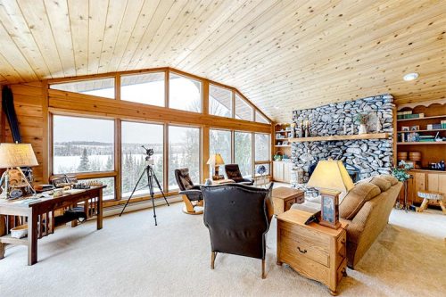 Cozy living room with wooden walls, large windows, and a stone fireplace, overlooking a snowy landscape.