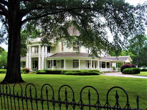 A large, elegant white house with a wraparound porch, surrounded by green grass and trees, viewed through a black fence.