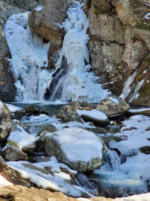 A frozen waterfall cascading over rocky terrain, surrounded by snow-covered boulders and icy patches.