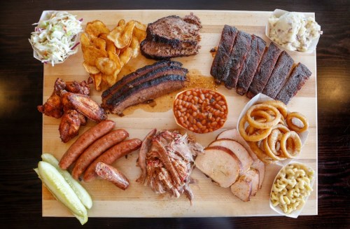 A wooden platter filled with various meats, sides like coleslaw, baked beans, onion rings, and pickles.