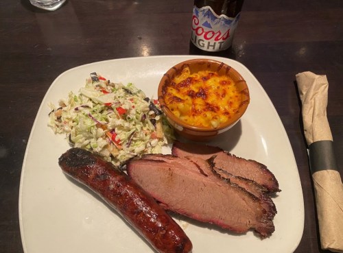 A plate with sliced brisket, a sausage, coleslaw, mac and cheese, and a Coors Light beer.