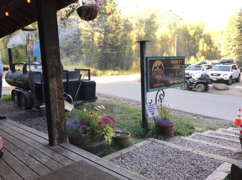 A rustic outdoor scene featuring a sign for a lodge, flower pots, and parked vehicles along a gravel path.
