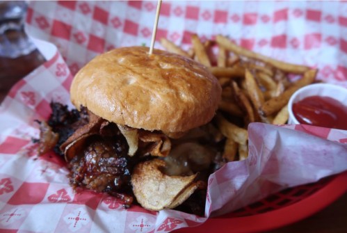 A hearty burger topped with bacon and chips, served with fries and a side of ketchup in a red-checkered basket.