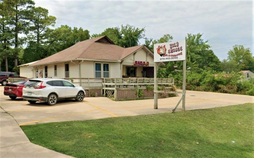 A small restaurant with a wooden deck, surrounded by trees, featuring a sign and parked cars in the lot.
