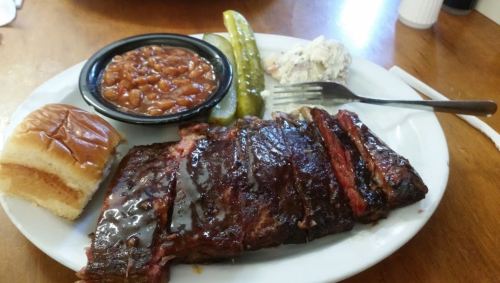 A plate featuring sliced barbecue ribs, baked beans, a pickle, coleslaw, and a bun on a wooden table.