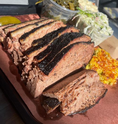 Sliced smoked brisket with a charred crust, served with sides of coleslaw and pickles on a tray.
