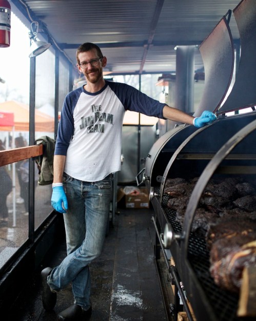 A man in a t-shirt and gloves stands beside a large smoker, grilling meat at an outdoor event.
