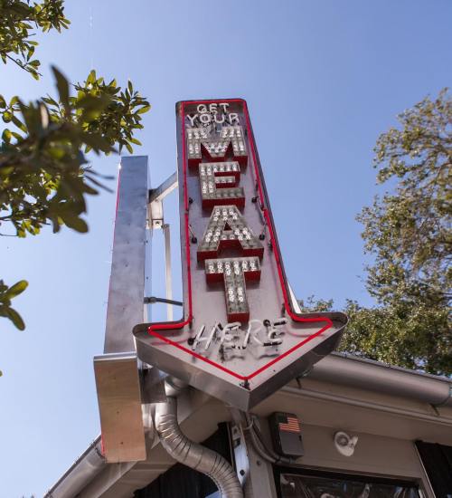 A retro-style sign reads "Get Your Meat Here" with bright lights, surrounded by trees and blue sky.