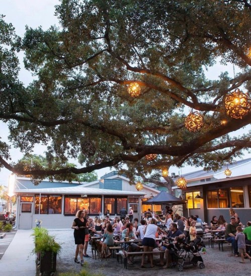 A lively outdoor dining area under a large tree, with people enjoying meals and decorative lanterns hanging above.