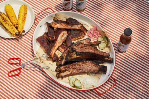 A platter of smoked meats, including ribs and brisket, served with pickles, onions, and corn on the side.