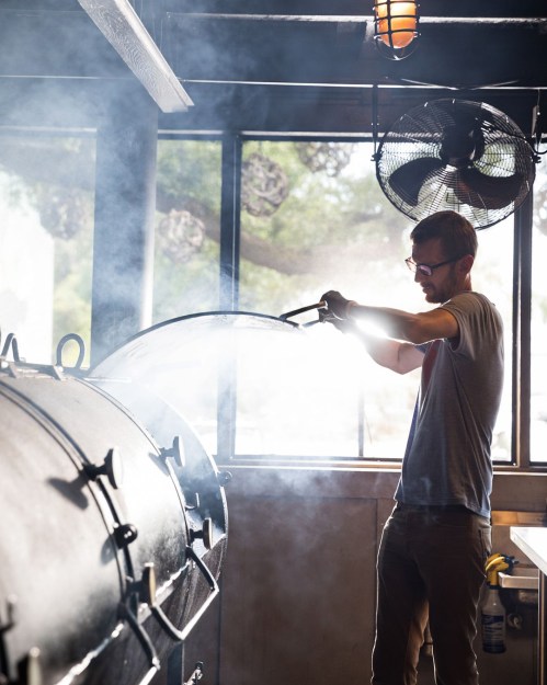 A person in a gray shirt tends to a large smoker, releasing steam in a well-lit, industrial kitchen setting.