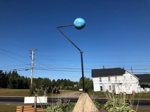 A blue sphere mounted on a metal arm, set against a clear blue sky, near a road and a white house.