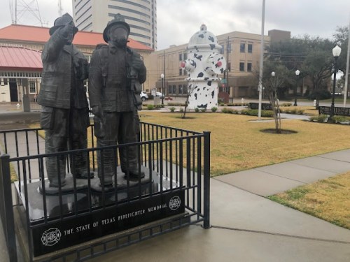 Statue of two firefighters at the Texas Firefighter Memorial, with a colorful sculpture in the background.