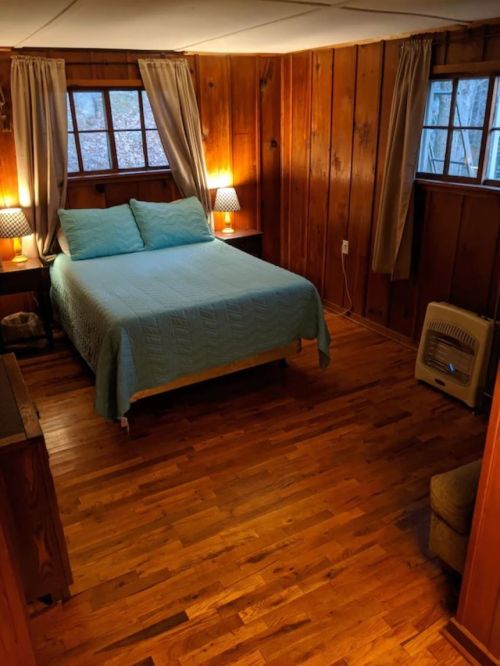 Cozy bedroom with wooden walls, a blue quilted bedspread, two lamps, and windows letting in natural light.