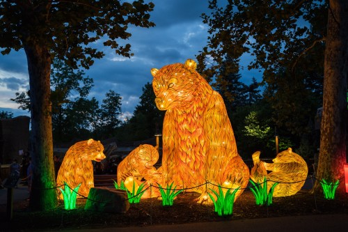 A glowing bear sculpture surrounded by smaller bear figures, illuminated against a twilight sky and greenery.