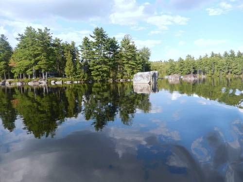 A serene lake scene with trees and rocks reflecting on the calm water under a partly cloudy sky.