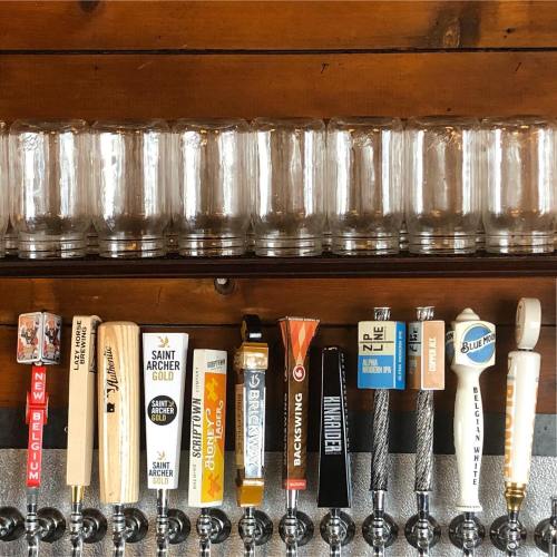 A row of beer taps with various labels and glasses on a wooden bar top.