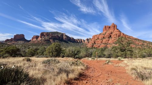 Red rock formations rise against a blue sky, surrounded by desert vegetation and a dirt path in the foreground.