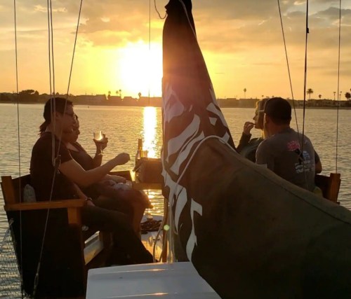 A group of people enjoying drinks on a boat at sunset, with a sail partially obscuring the view.