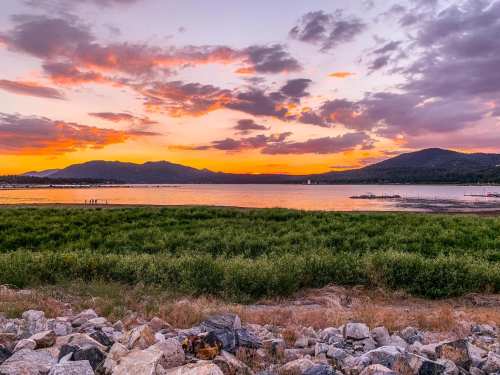 A vibrant sunset over a lake, with mountains in the background and rocky foreground, creating a serene landscape.