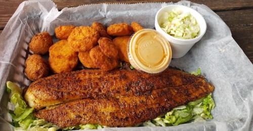 A platter featuring a seasoned fish fillet, golden fried nuggets, coleslaw, and a dipping sauce on a bed of lettuce.