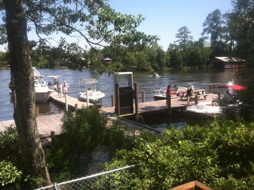A sunny riverside scene with boats docked, people enjoying the water, and lush greenery surrounding the area.