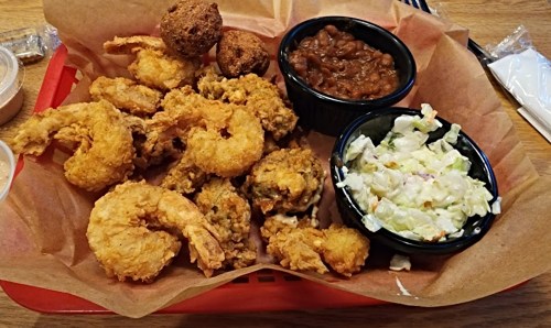 A tray of fried shrimp, hushpuppies, coleslaw, and baked beans served on brown paper.
