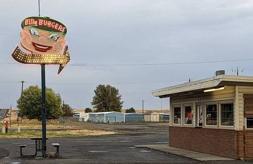 A retro burger joint sign featuring a smiling cartoon chef, with a small building nearby and a cloudy sky.