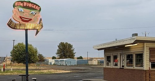 A retro burger joint sign with a smiling face, next to a small building in a rural area under a cloudy sky.