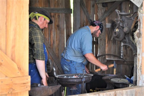 Two blacksmiths working in a rustic workshop, one hammering metal while the other observes. Tools and equipment are visible.