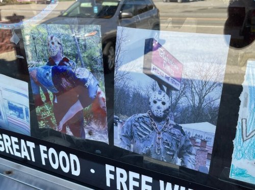 Two posters of a masked figure displayed in a window, with a sign for a restaurant in the background.