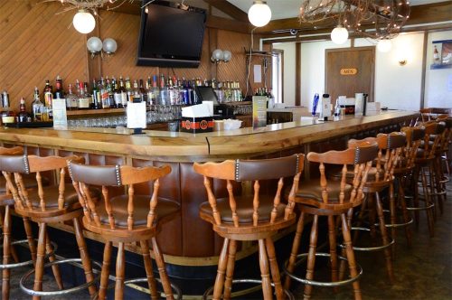A wooden bar with high stools, a well-stocked liquor shelf, and a large TV on the wall in a cozy setting.