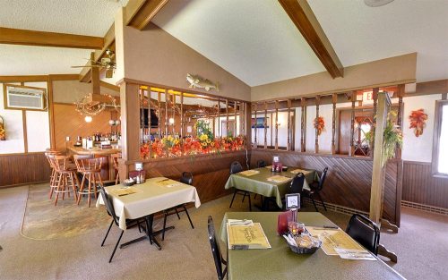 Cozy restaurant interior with wooden beams, tables set for dining, and autumn decorations. Bar area in the background.