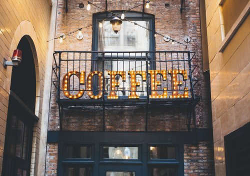 A rustic alleyway with a balcony displaying a bright, illuminated "COFFEE" sign.