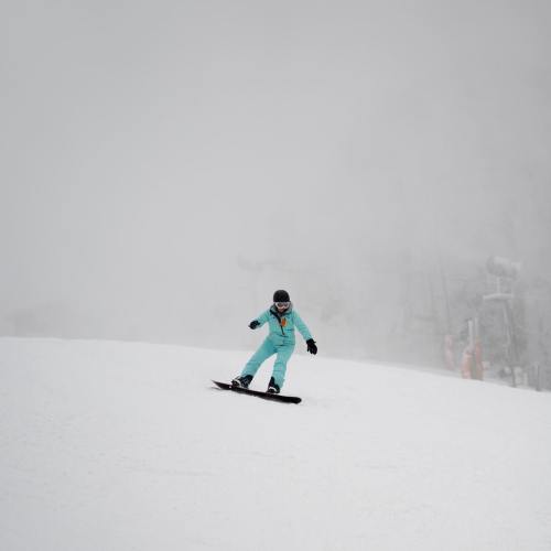 A snowboarder in a teal outfit glides down a snowy slope amidst foggy weather.