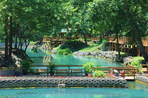 A serene park scene featuring a calm blue waterway, lush greenery, wooden walkways, and colorful flower beds.
