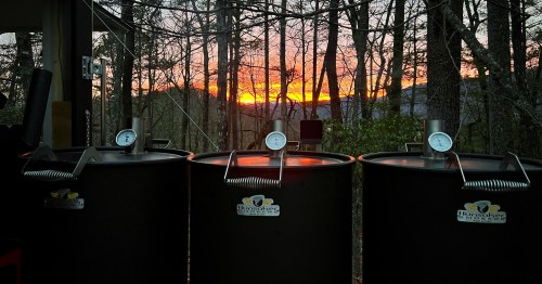Three black barrels with gauges sit in front of a vibrant sunset, framed by trees in a serene outdoor setting.