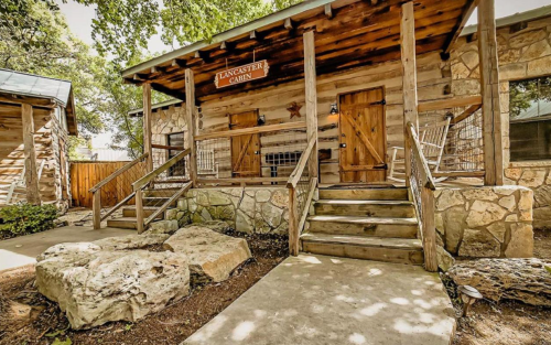 A rustic cabin entrance with wooden steps, a sign reading "Lancaster Cabin," and stone landscaping.