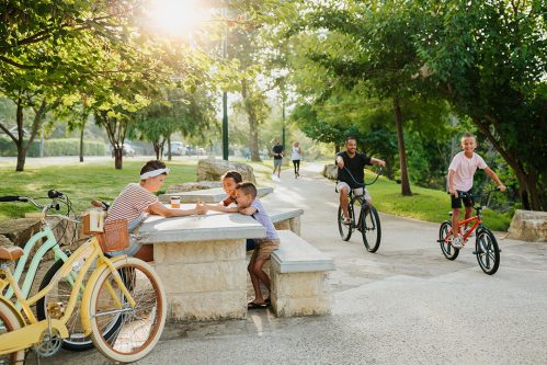 A sunny park scene with children playing, adults biking, and a group enjoying drinks at a stone table.