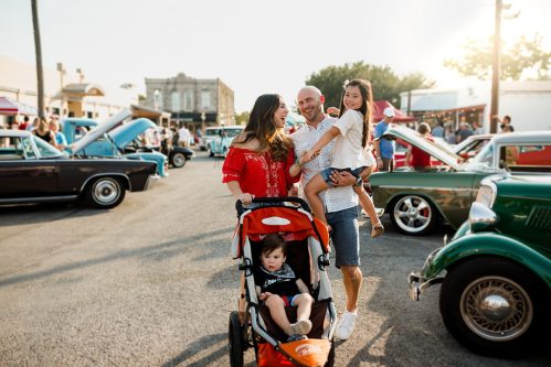 A family enjoys a car show, with a woman pushing a stroller, a man holding a girl, and classic cars in the background.