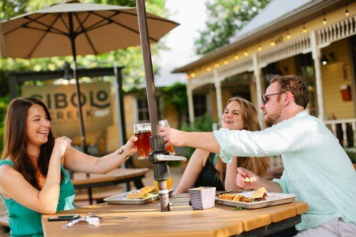 Three friends toast with drinks at an outdoor table, enjoying food and laughter in a lively setting.