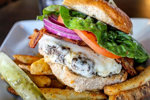 A close-up of a juicy burger with lettuce, tomato, onion, and cheese, served with crispy fries and a pickle on the side.