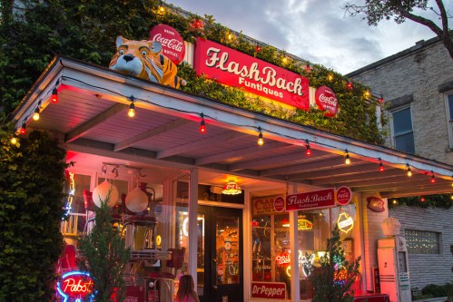 A vibrant storefront of FlashBack Funtiques, adorned with neon signs and colorful lights, surrounded by greenery.