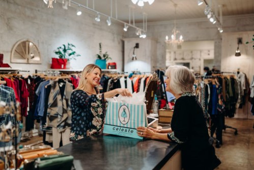A smiling customer receives a shopping bag from a cashier in a stylish boutique filled with clothing.