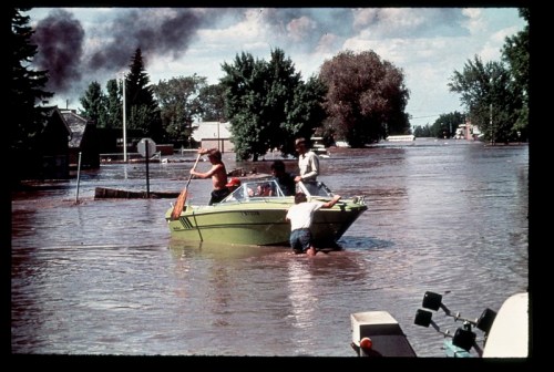 A group of people in a boat navigating through a flooded neighborhood, with submerged houses and trees in the background.