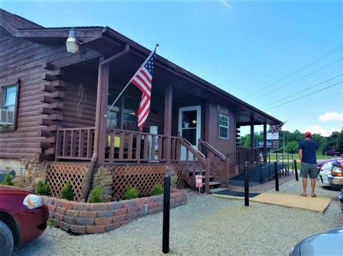 A wooden building with an American flag, featuring a ramp and a gravel parking area, under a clear blue sky.