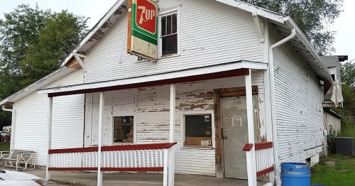 Abandoned white house with a faded 7UP sign, peeling paint, and a blue barrel in front.